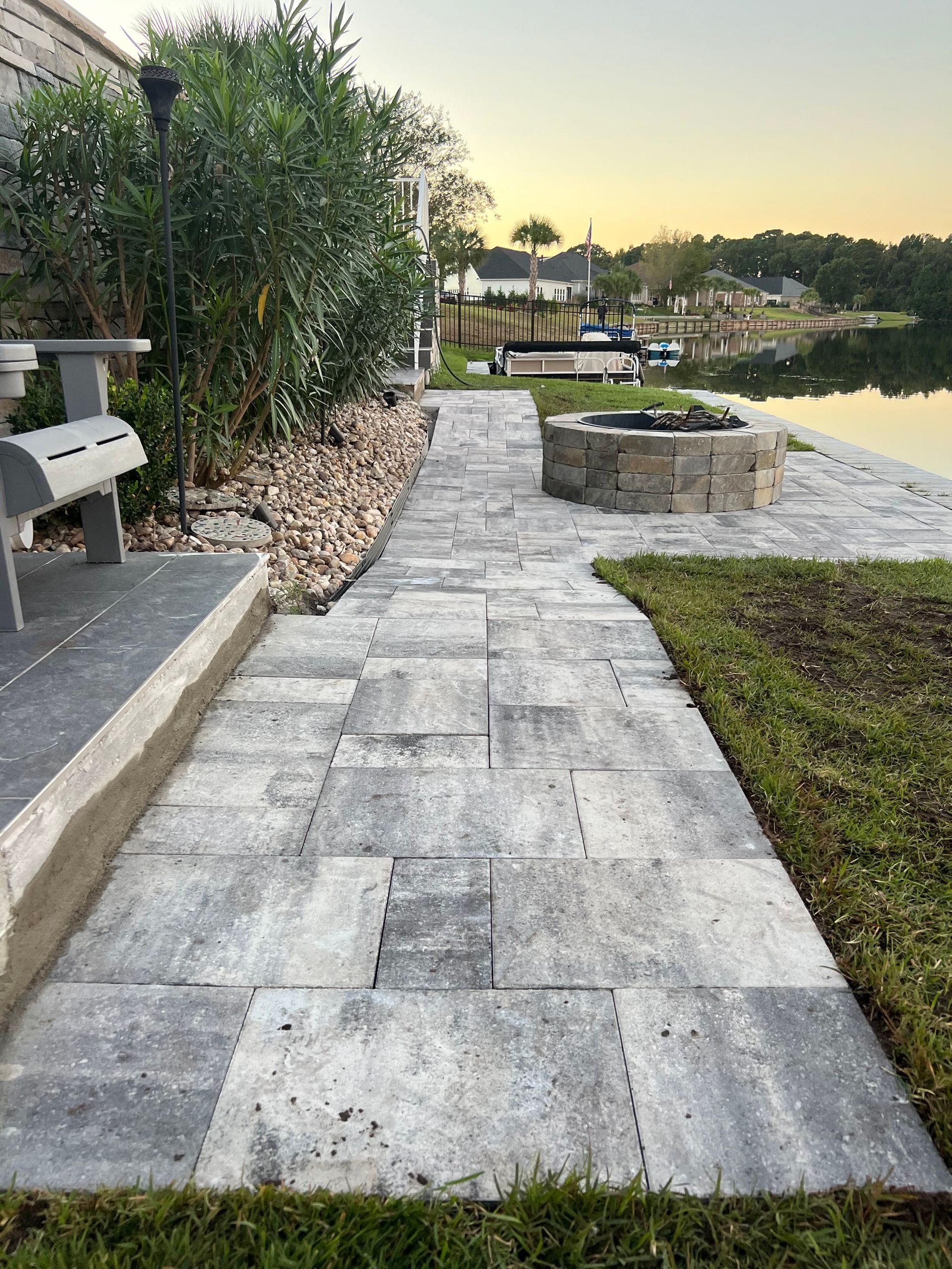 Stone walkway beside water and greenery at sunset, with a bench and fire pit area.