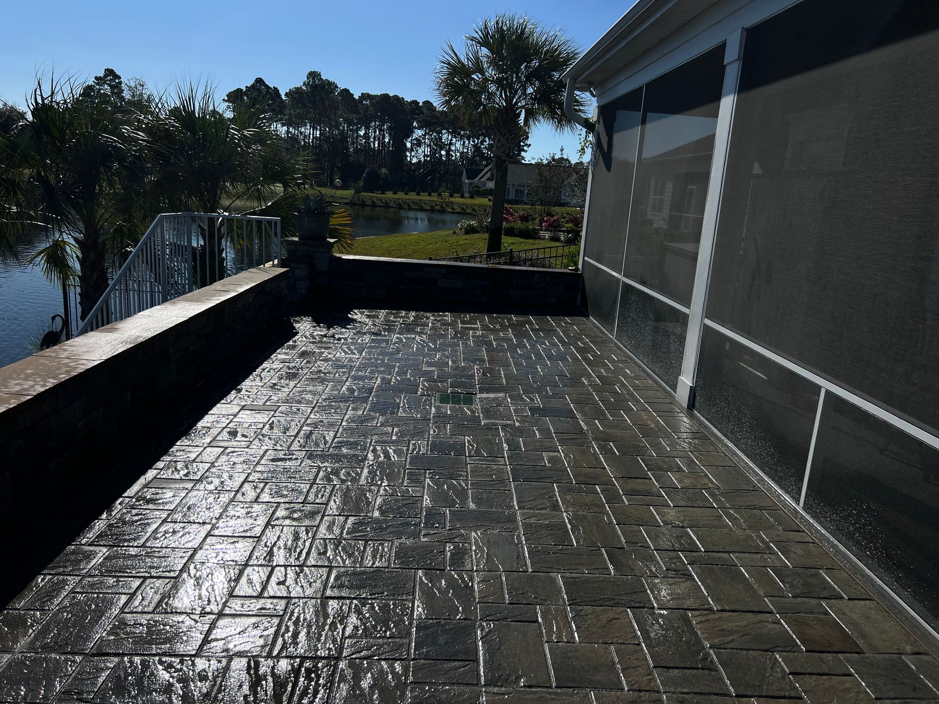Sunlit brick patio beside a house with a low wall and fenced yard, shadows across the wet surface.