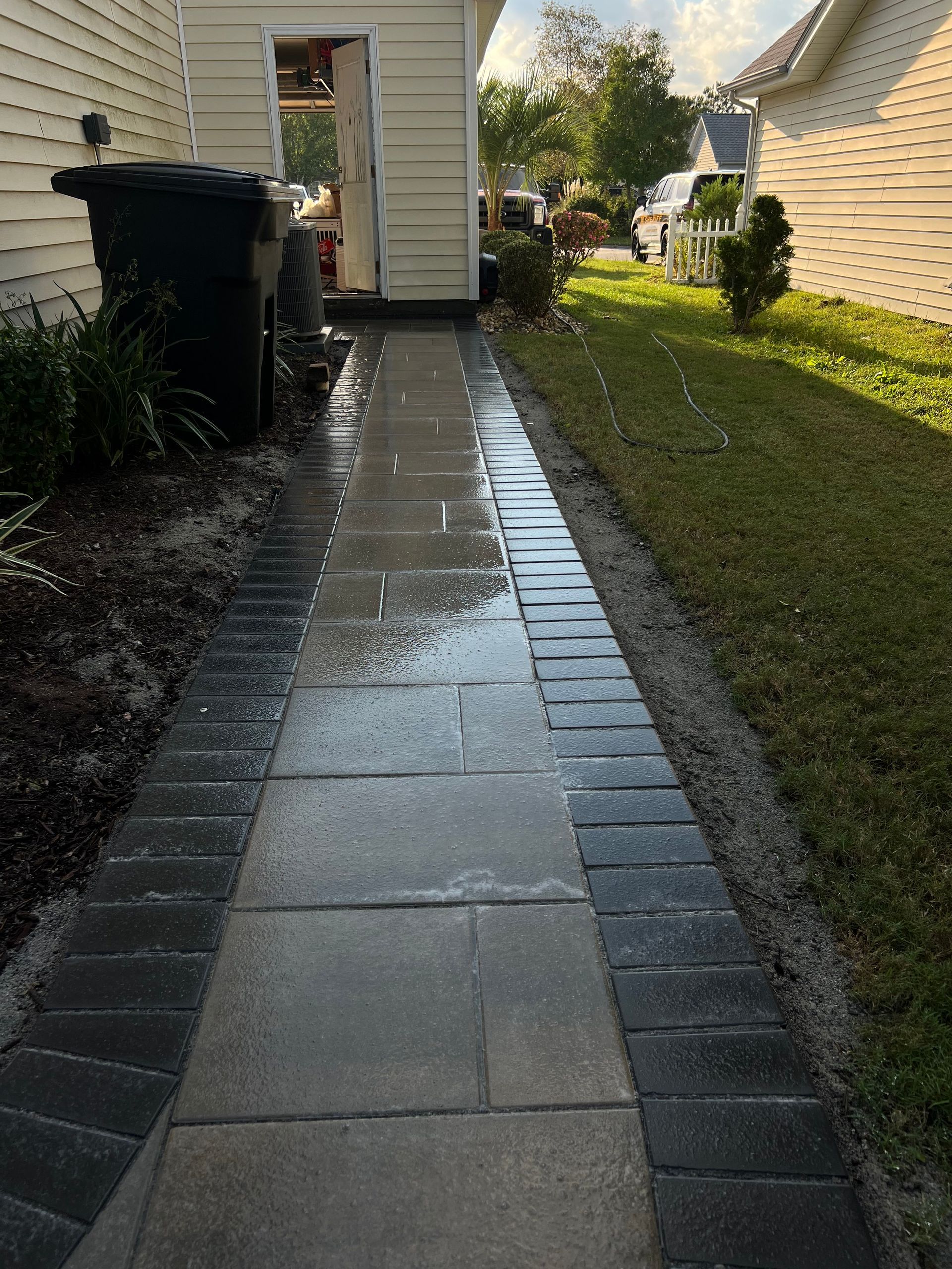 Wet stone walkway beside a house, bordered by brick and grass after rain.