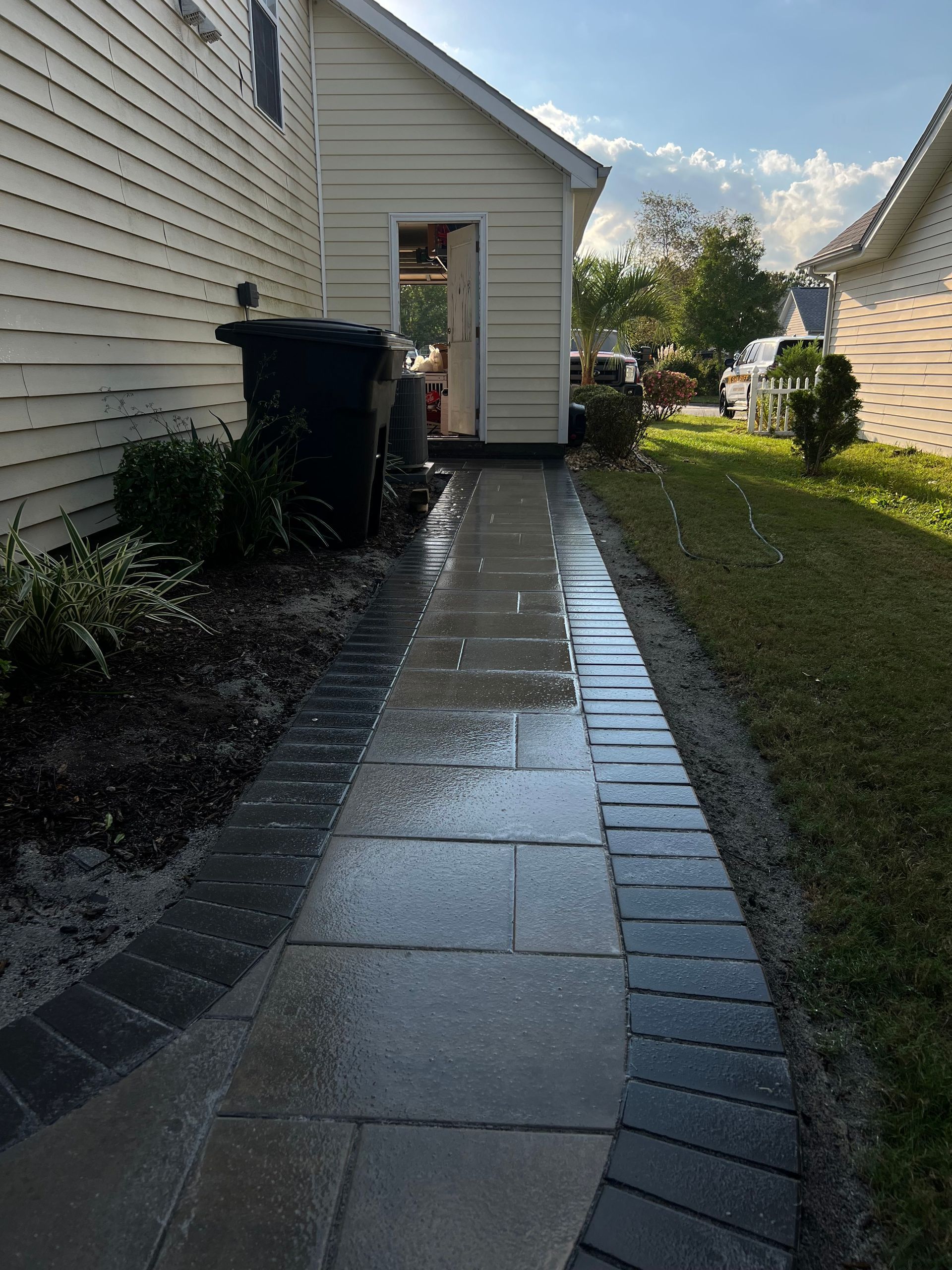 Wet stone walkway beside a white house, leading past a side door into a sunny yard.