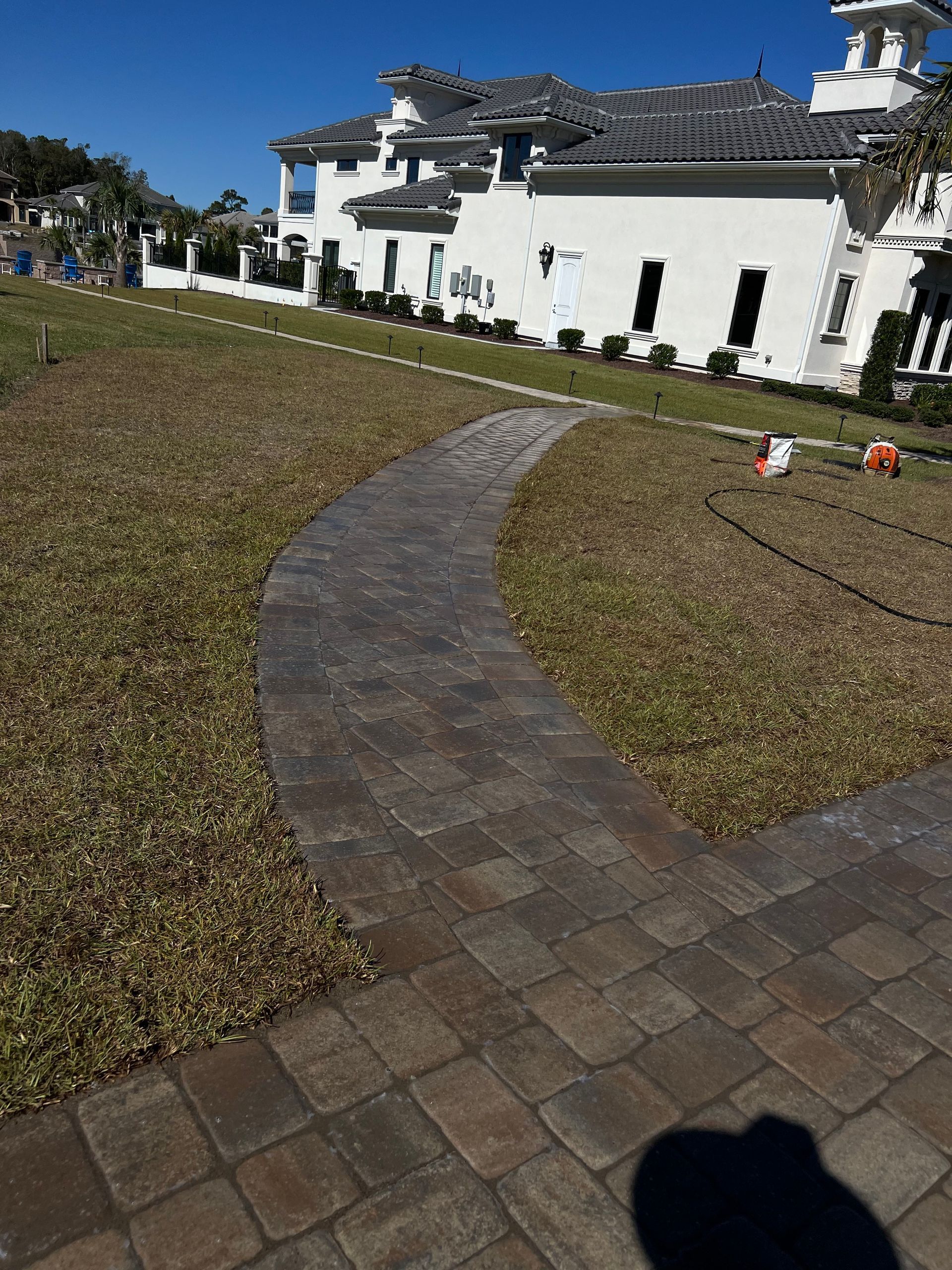 Curved brick path through a grassy lawn toward a white house with a pergola in bright sunlight