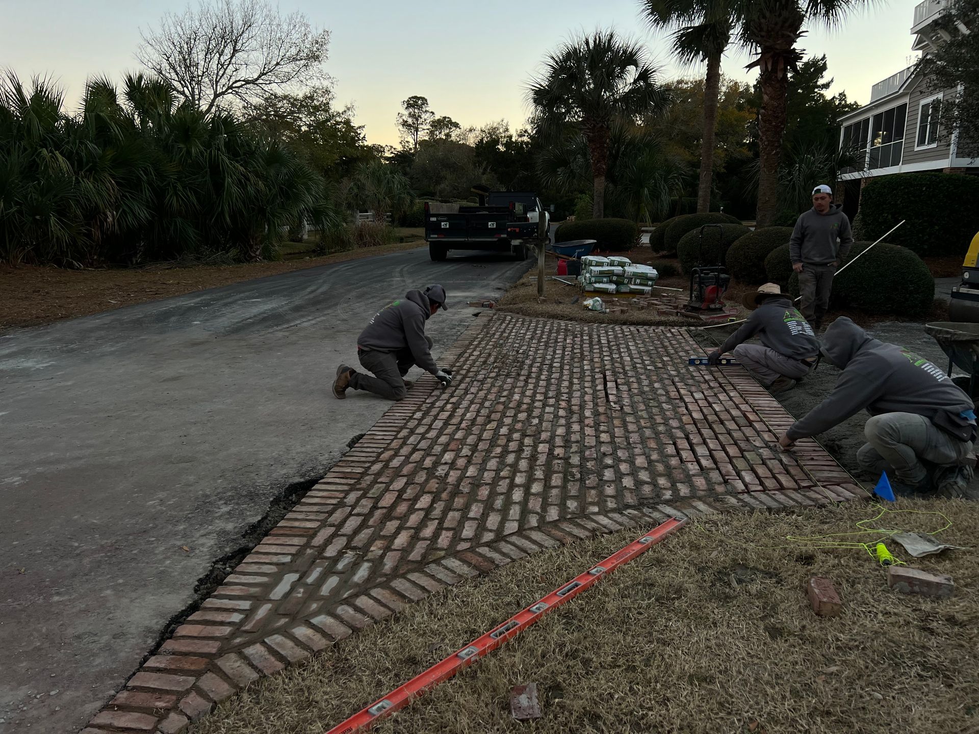 Workers laying brick pavers on a driveway beside a road at dusk