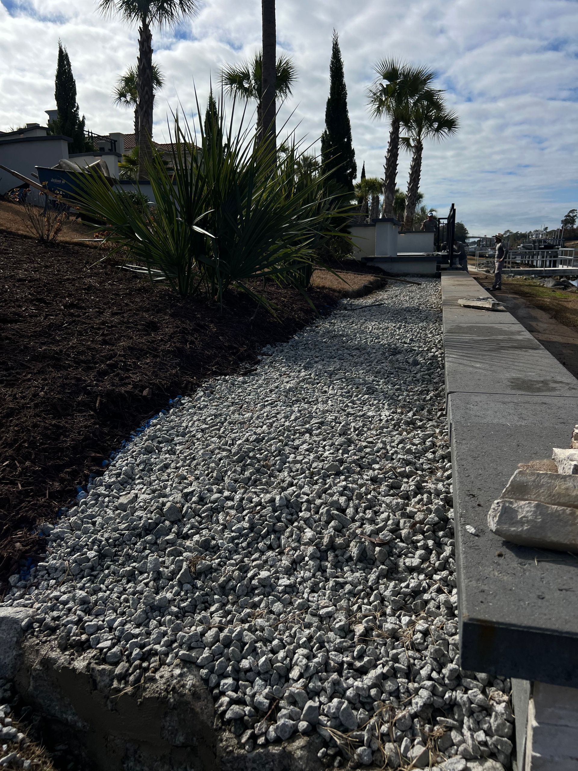 Gravel path beside a landscaped hillside with shrubs, tall cypress trees, and a bright sky beyond the overlook.