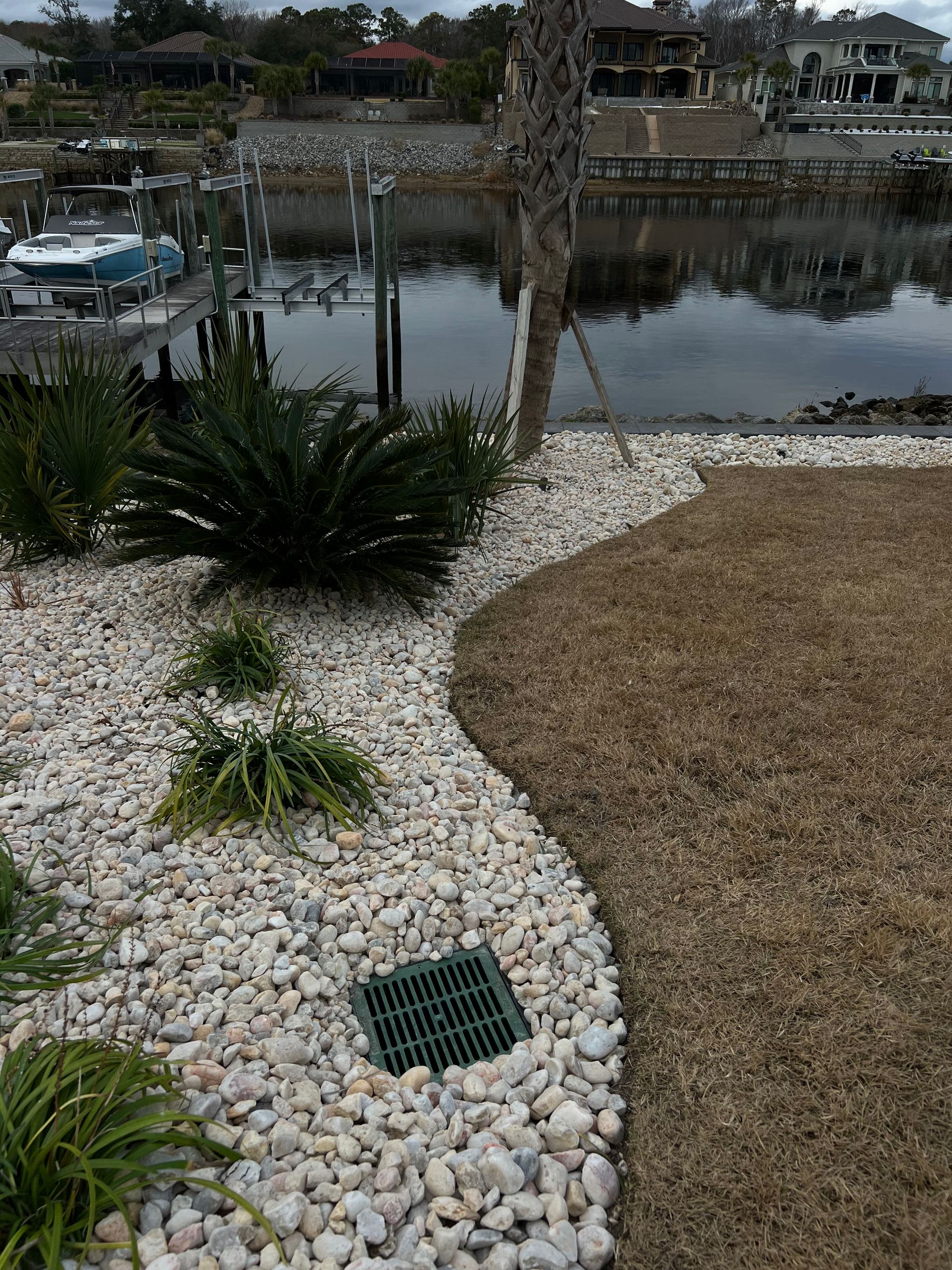 Curved white-rock garden border beside a brown lawn, with a waterfront and moored boats in the background