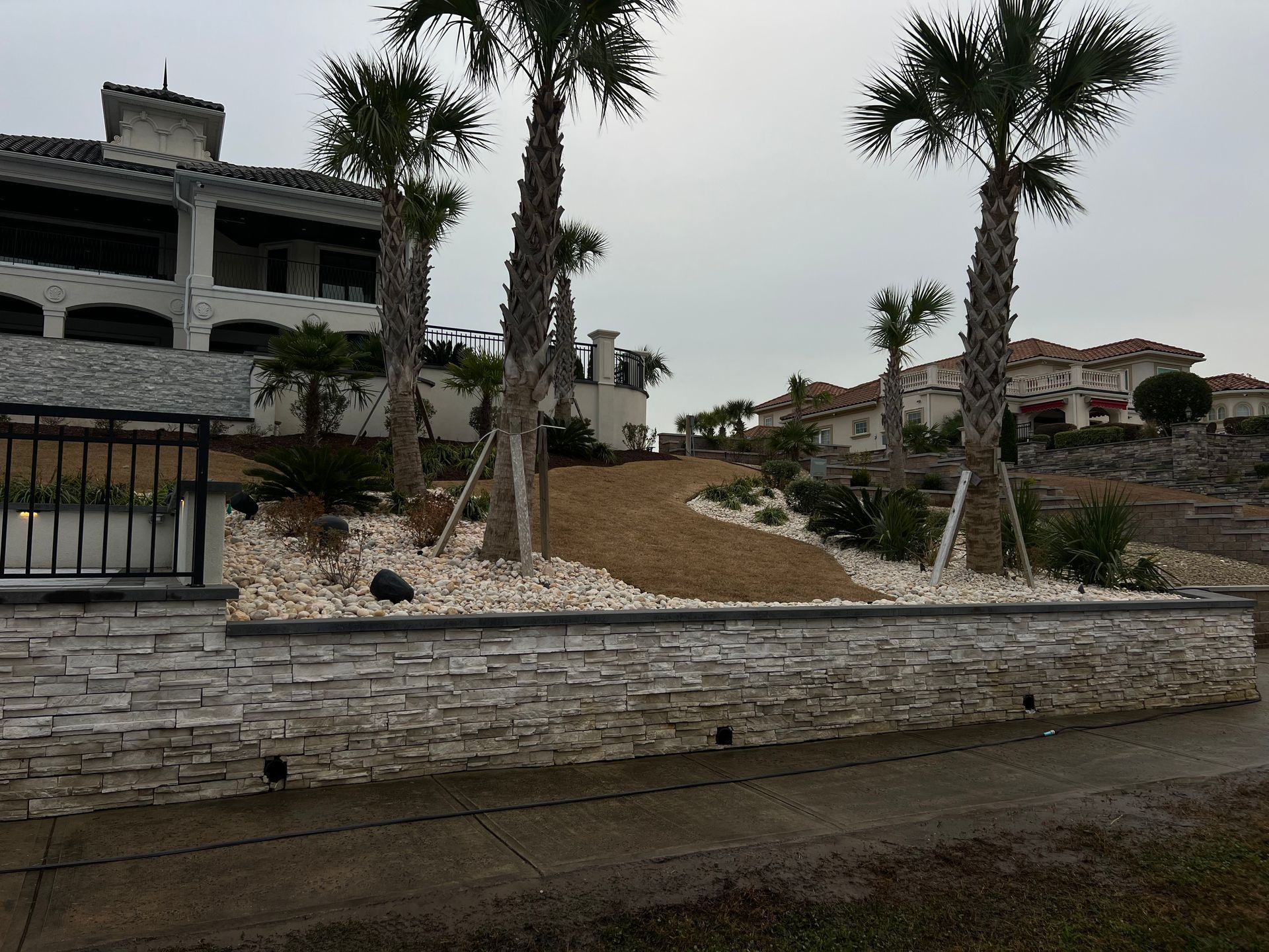 Landscaped hillside with palm trees and stone retaining walls beside a large unfinished building