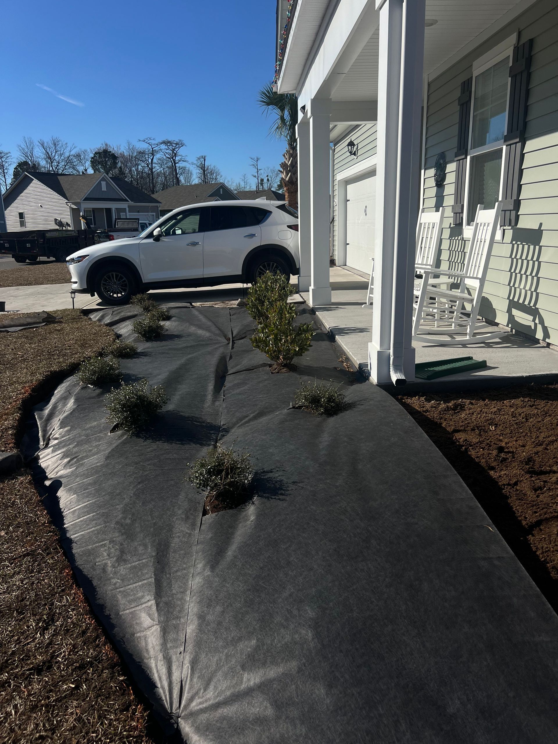 Front porch with black landscape fabric and small shrubs; white house and parked SUV in the background