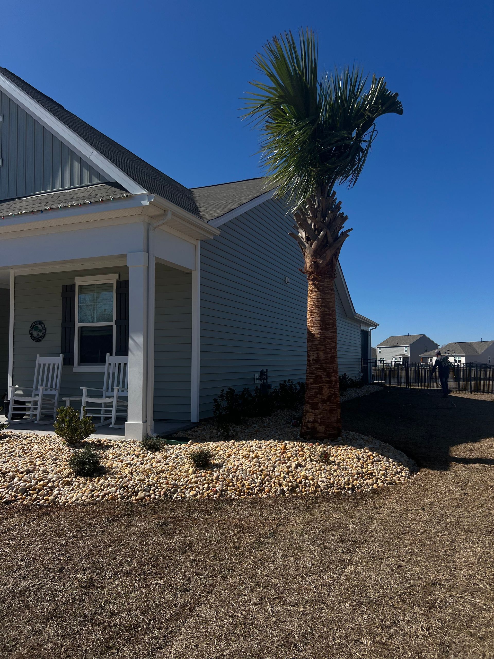 House exterior with white siding, front porch, and a palm tree in a gravel yard under a clear blue sky