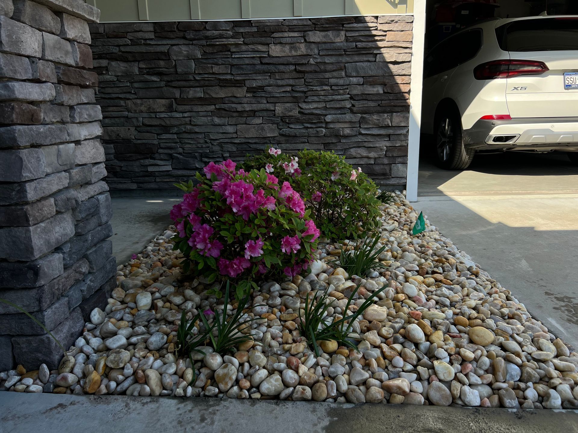 Pink flowers in a pebble rock garden beside a stone wall and parked white car