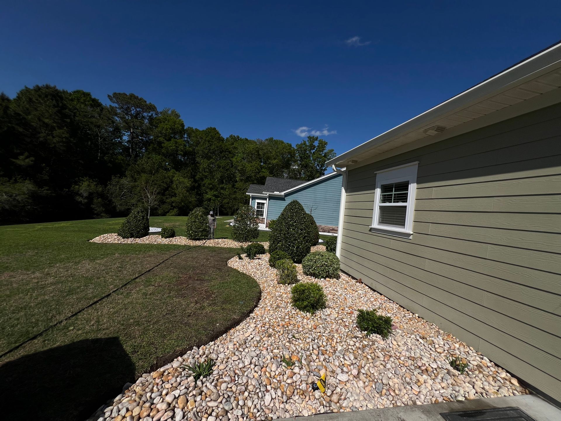Side yard with white gravel, trimmed shrubs, green lawn, and a beige house under a blue sky.