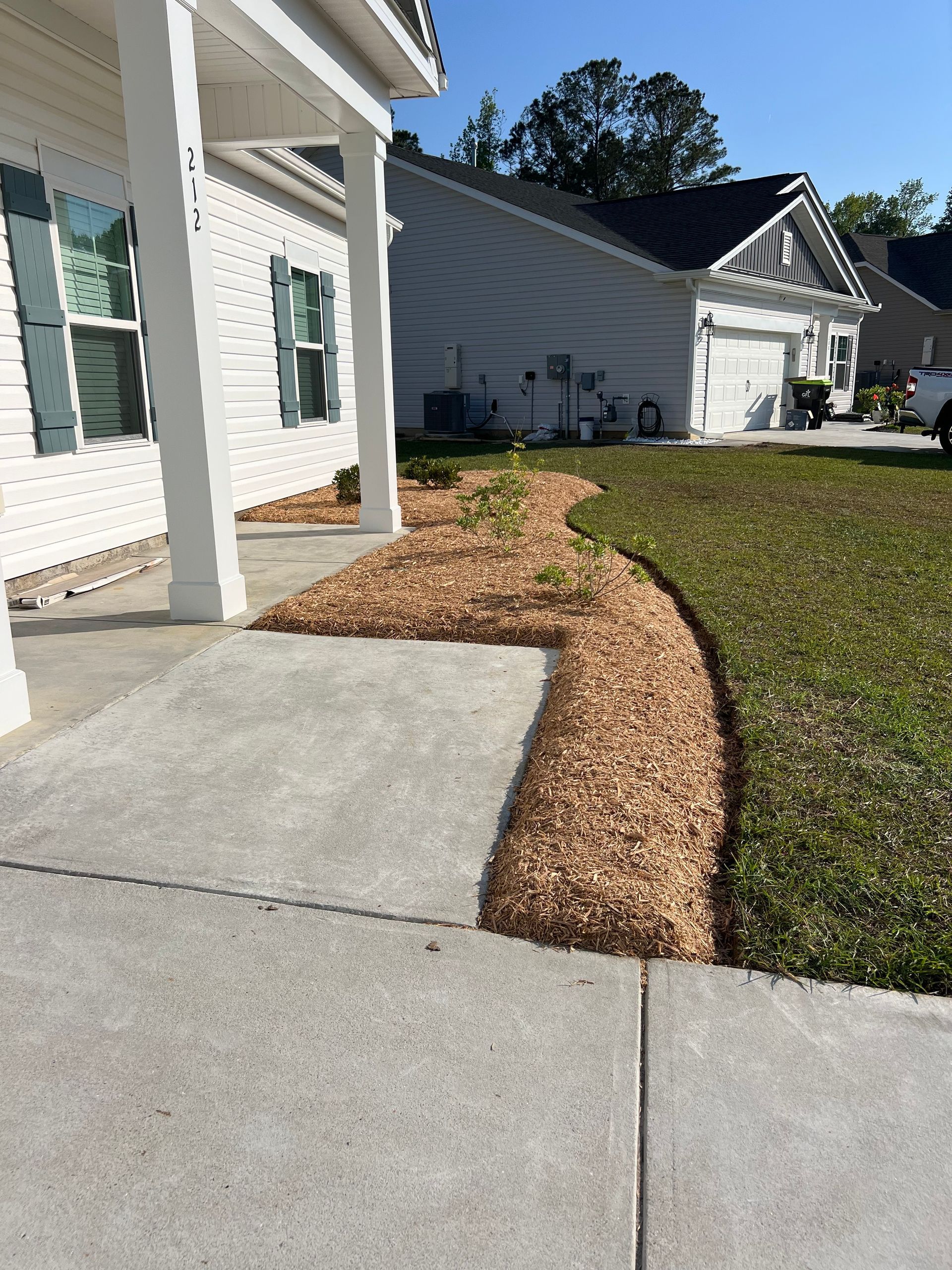 Concrete walkway beside a house with a narrow mulched border and green lawn in a suburban yard