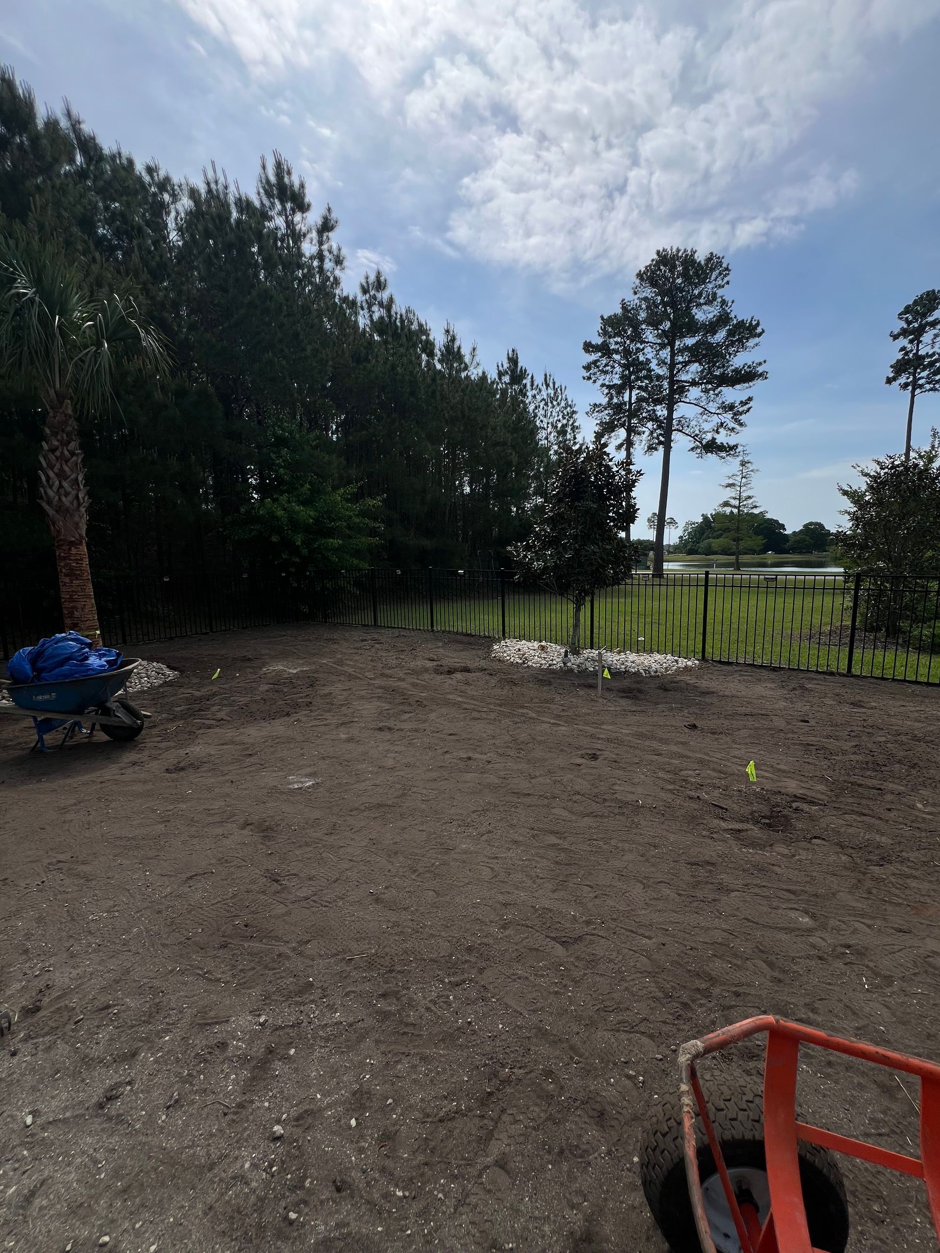 Dirt yard beside trees and a fence, with a cloudy sky overhead and a blue tarp-covered item at left.