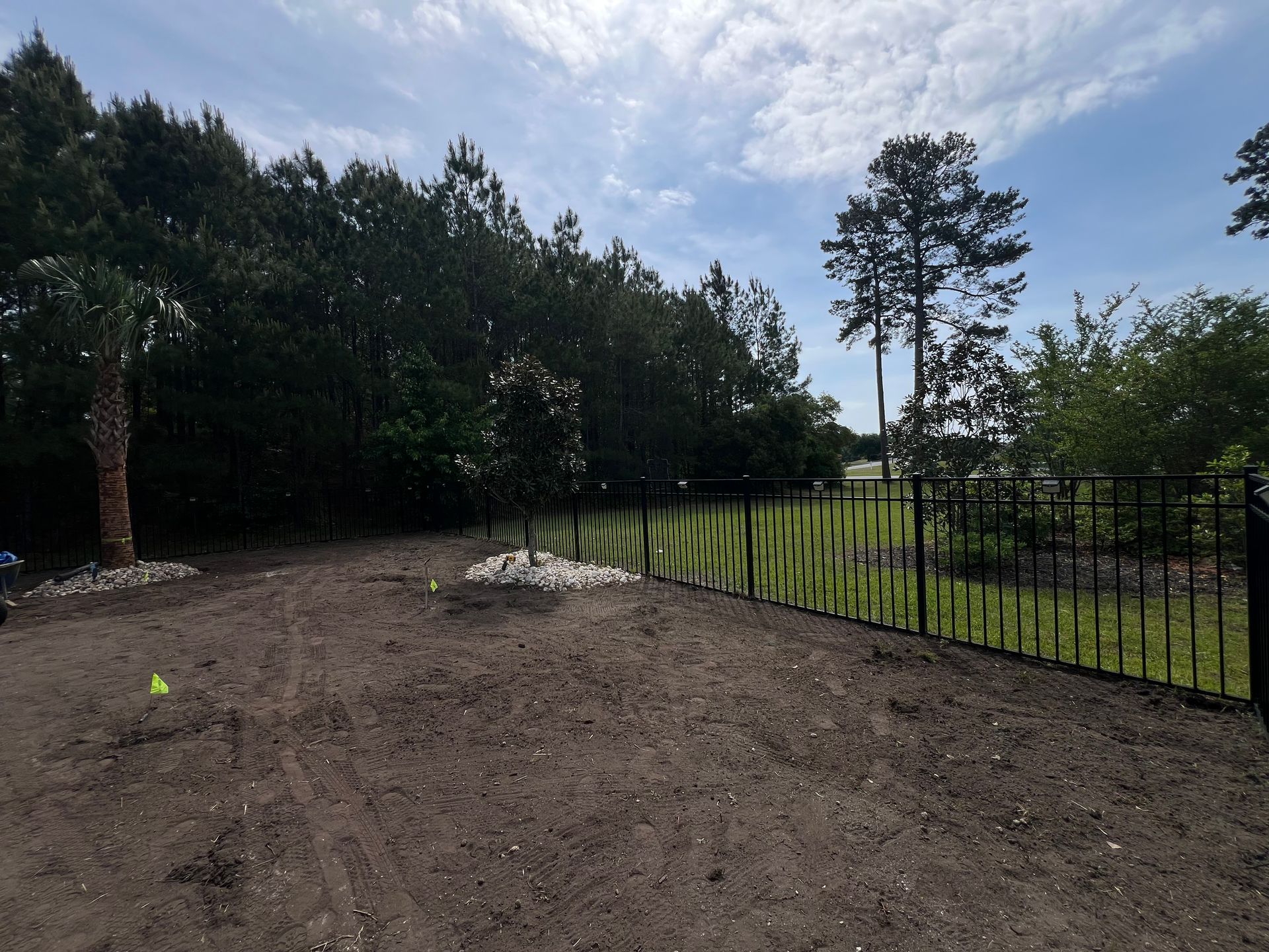 Freshly graded dirt yard beside a black metal fence, with trees and cloudy sky in the background