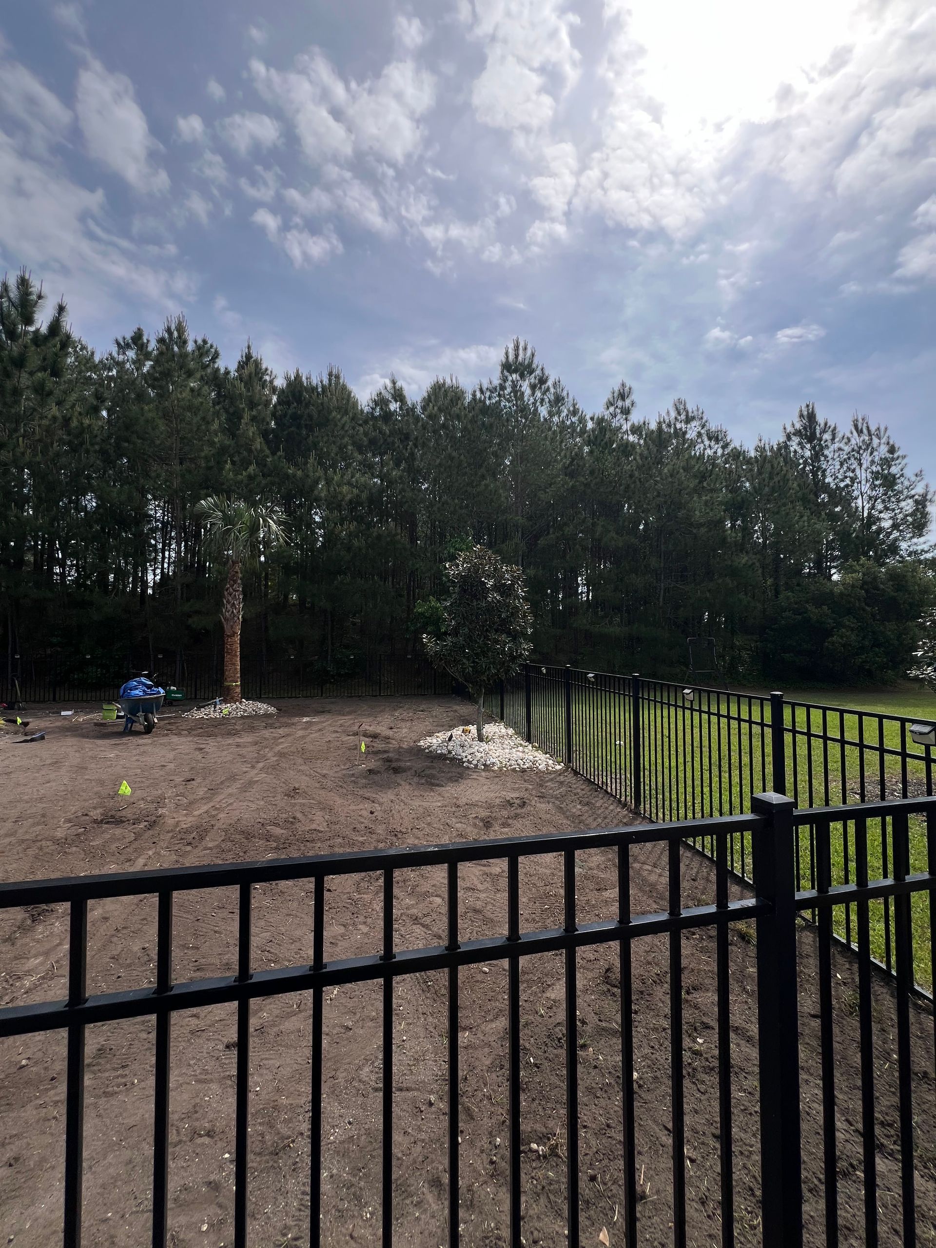 Black metal fence overlooking a gravel yard, trees, and cloudy sky beyond a grassy field.