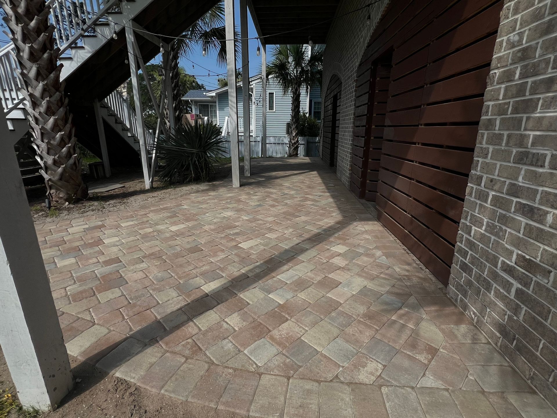 Brick-paved patio beside a house with a wooden wall, stairs, and palm trees in the background