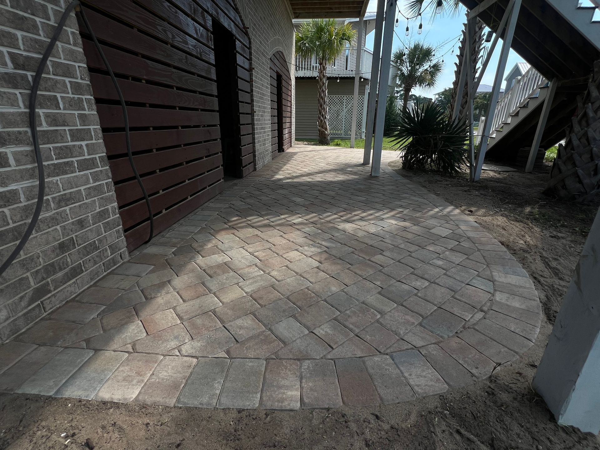 Brick-paved walkway beside a house, with shaded siding and nearby trees and plants.