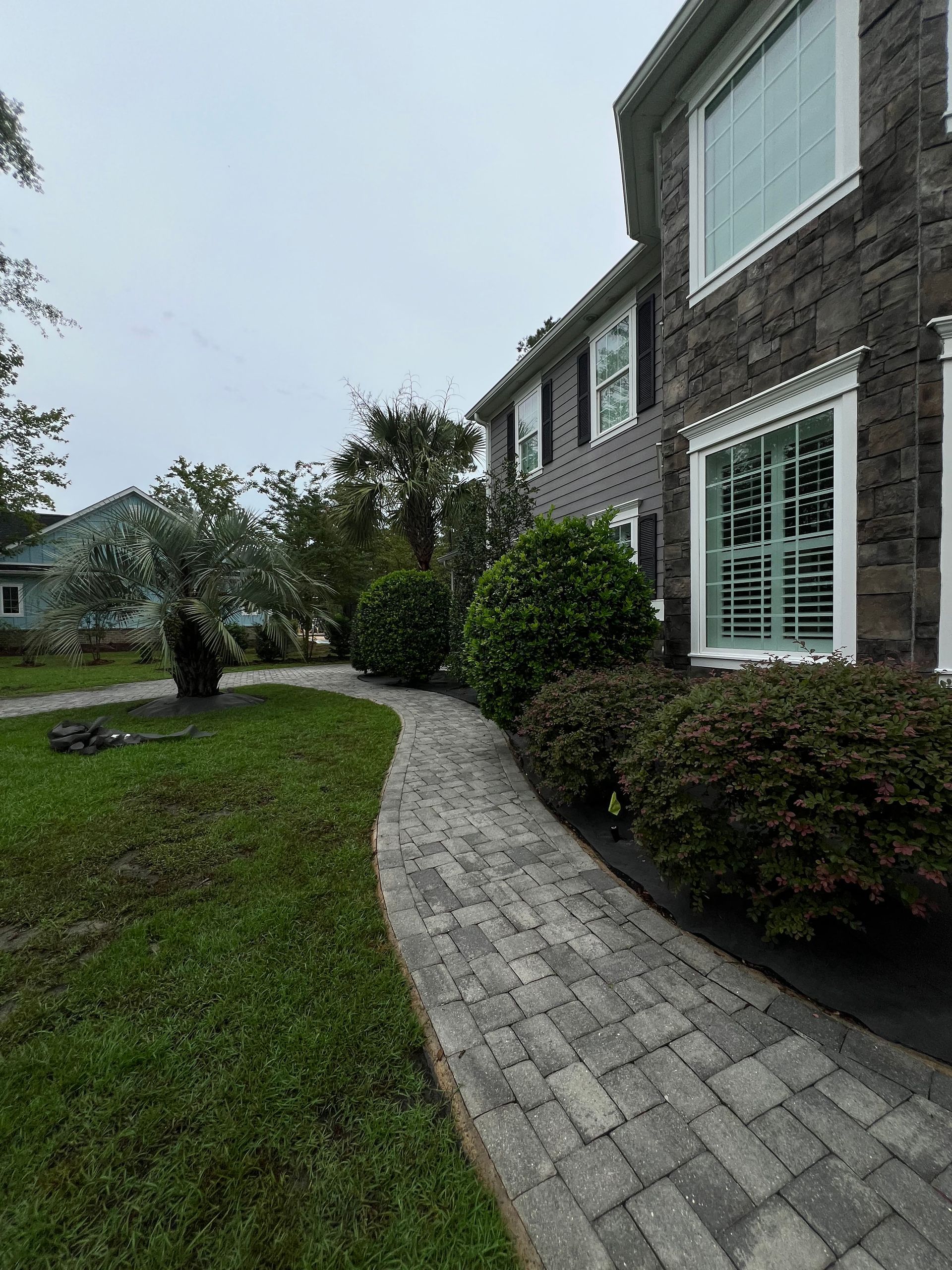 Curved brick walkway beside a two-story house with white-trimmed windows and green lawn on a cloudy day