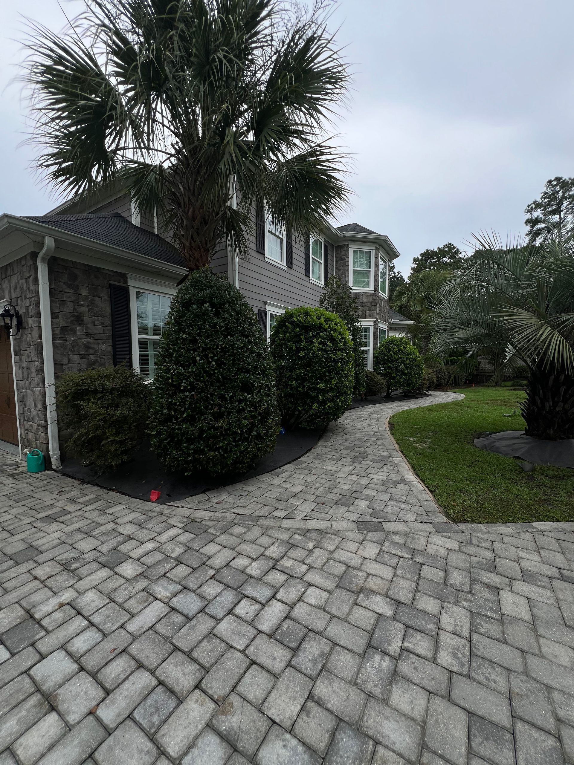 Stone-paver driveway and walkway leading to a gray house with palm trees and trimmed shrubs under cloudy skies