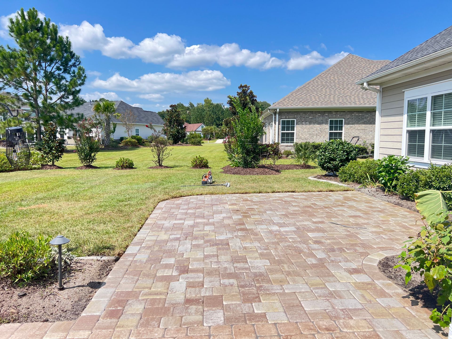 Brick patio beside a grassy suburban yard with shrubs, trees, and a house on the right.