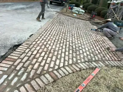 Workers laying brick pavers on a curved walkway beside grass and a driveway