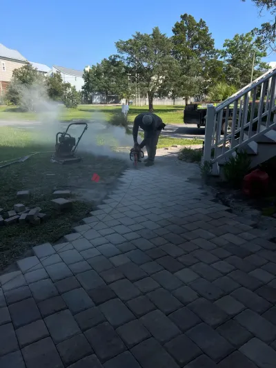 Person using a pressure washer on a brick walkway beside a house, with water spraying nearby.