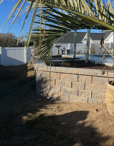 Backyard patio with a stone retaining wall, white fence, and palm fronds in the foreground.
