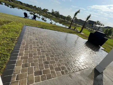 Sunlit brick patio beside a lake, with two people sitting on the grass and trees in the background