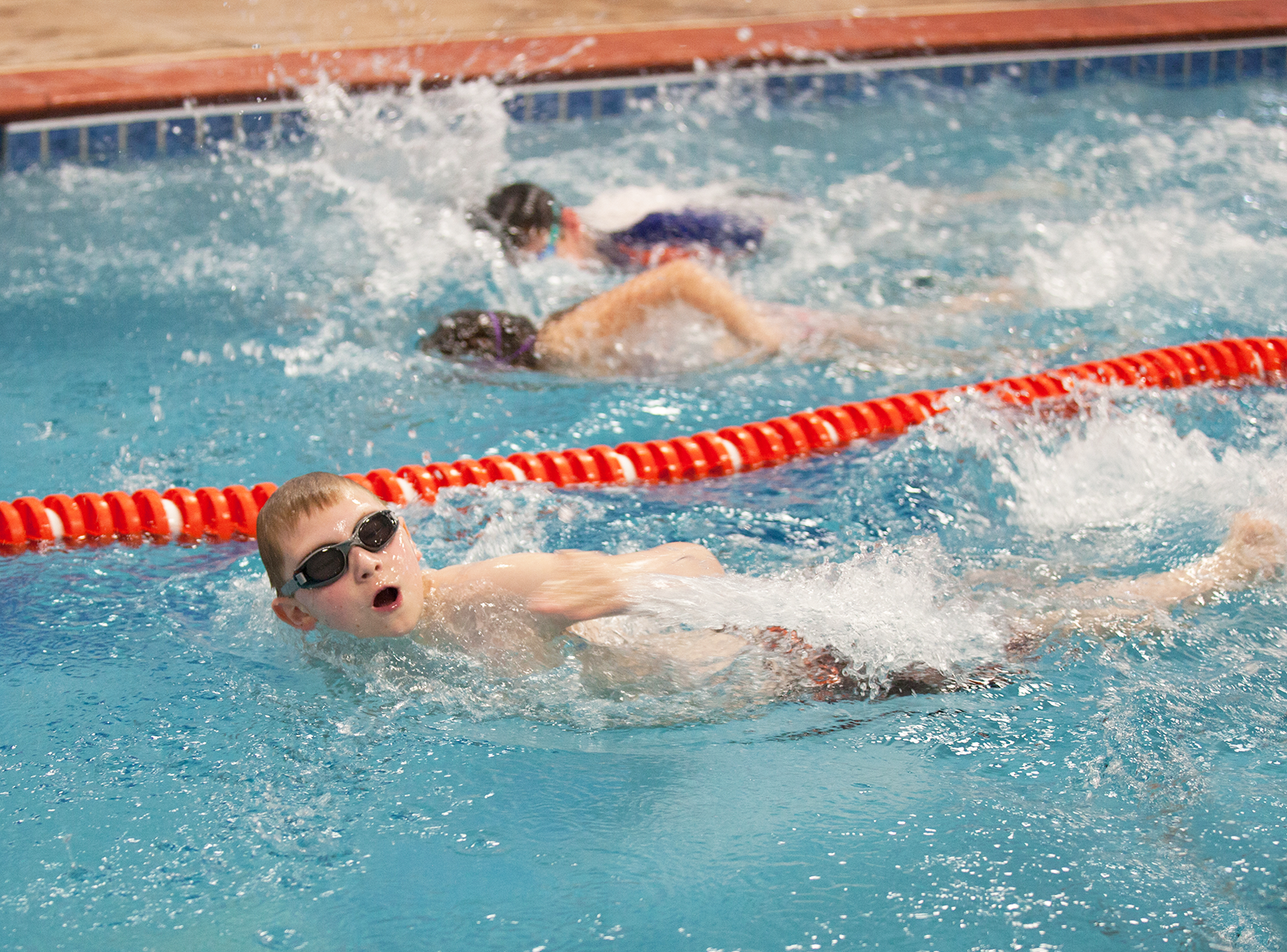 Swimmers in pool, performing the butterfly stroke. Water splashing, focused on the lead swimmer.