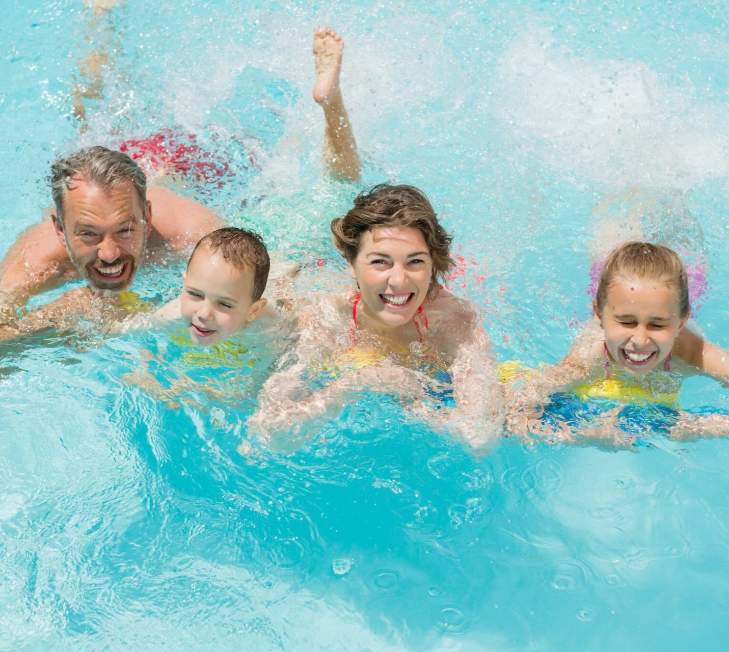 Family of four swimming in a clear blue pool, smiling and splashing.