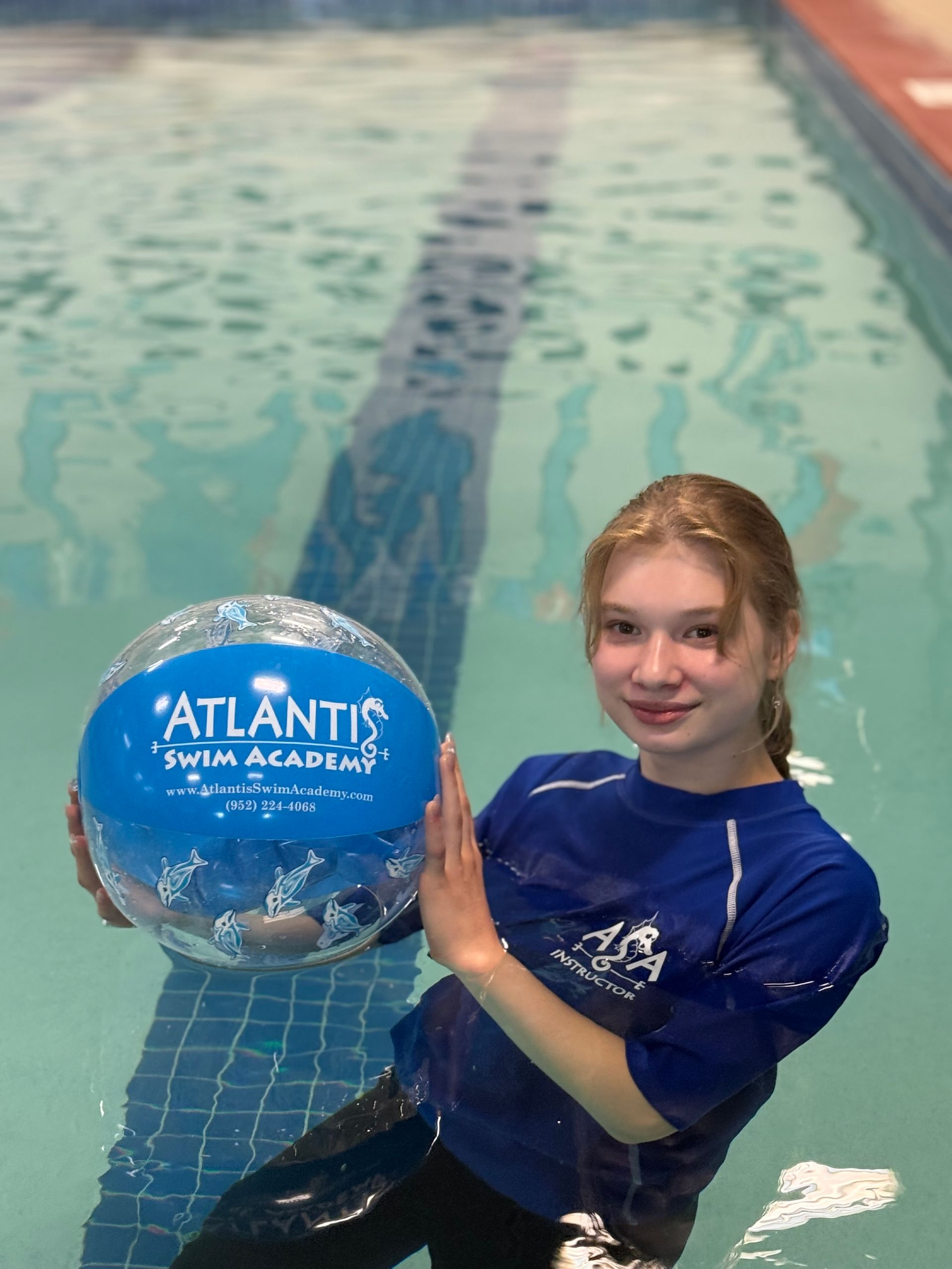 Woman smiling in pool, resting arms on black flotation device, wearing blue shirt.