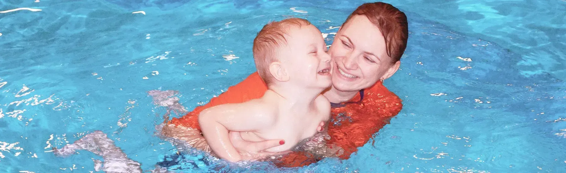 A baby being gently supported in a pool during a swim lesson with a smiling instructor.
