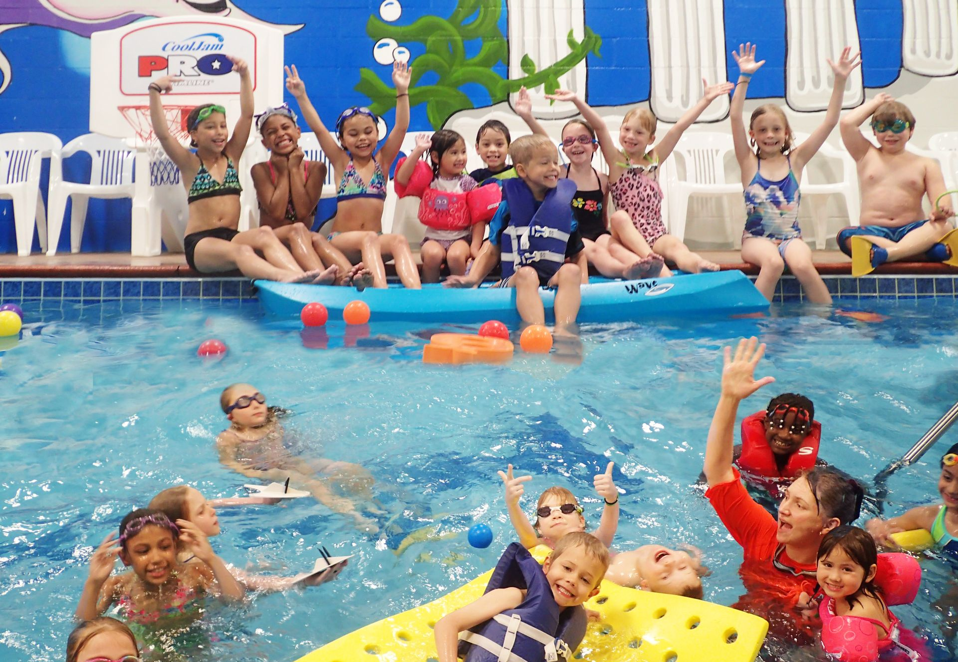 Children in a pool with arms raised. Some wear swimsuits and life vests. Poolside has chairs.