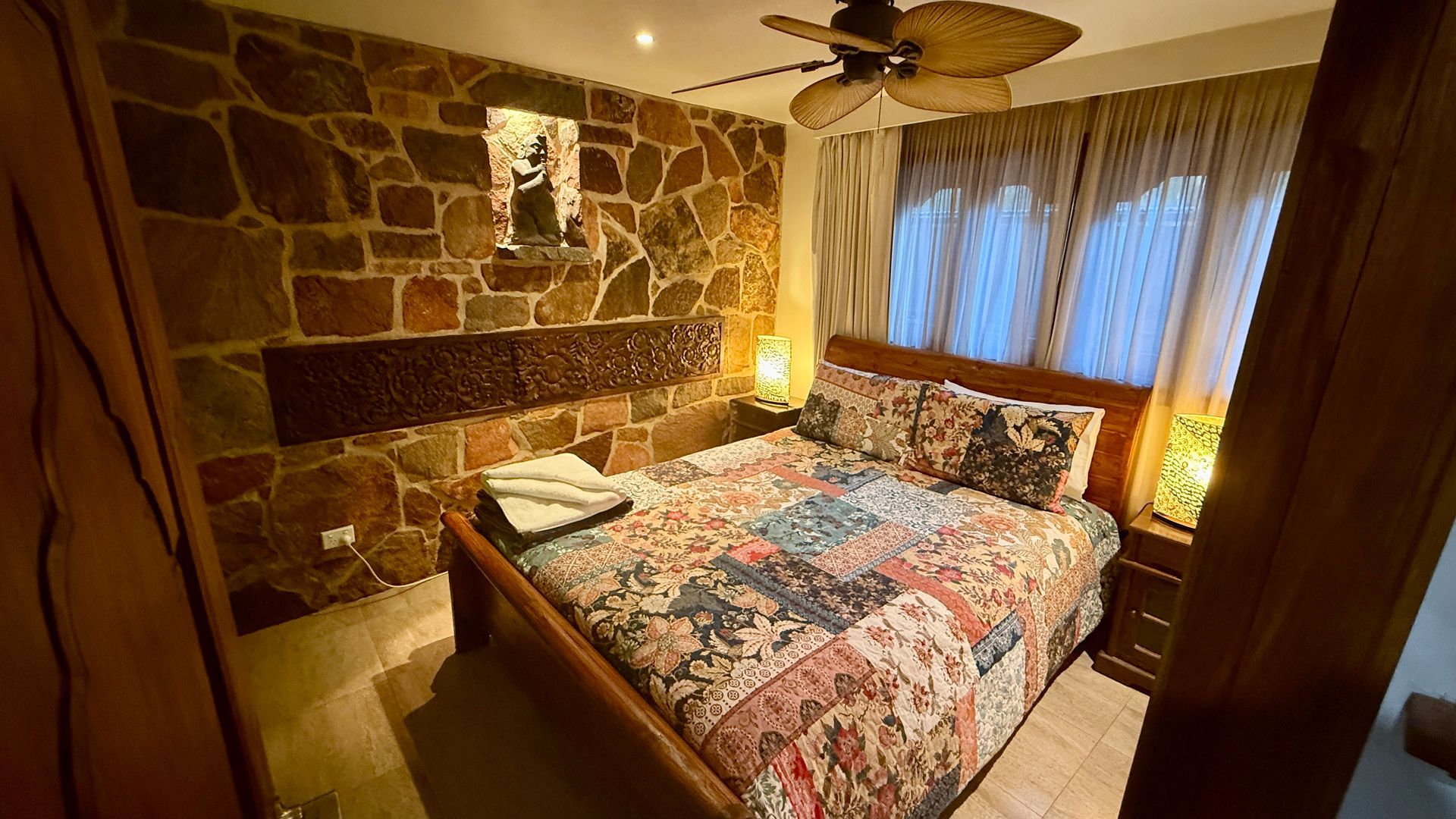 Bedroom with stone wall, bed, nightstand, and ceiling fan — Bali at the Bay in Nelson Bay, NSW