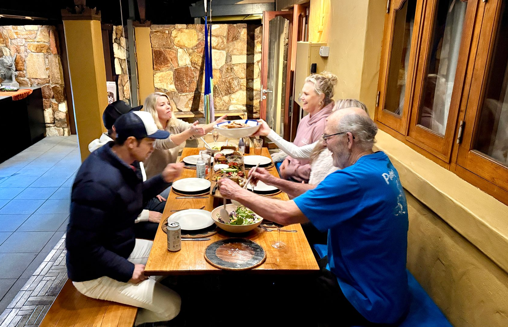 People at a wooden table, eating a meal together inside. They are passing plates.