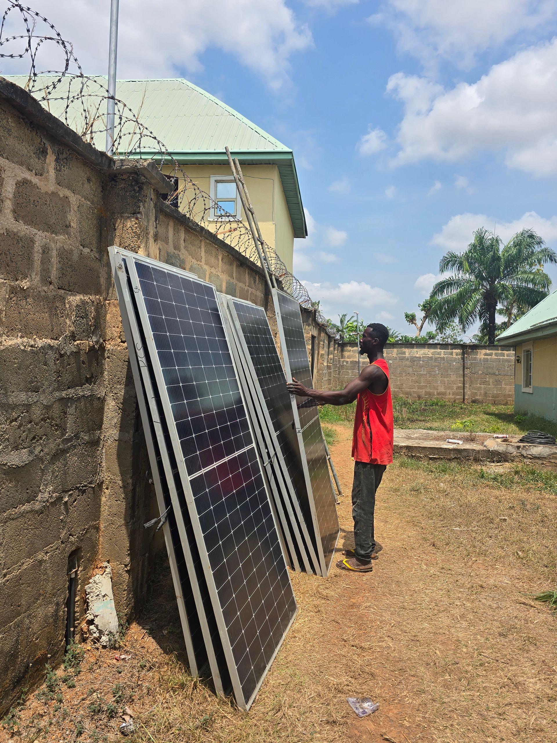 A man in a red shirt stands beside several solar panels propped against a brick wall topped with barbed wire, near a residential building with a green metal roof.