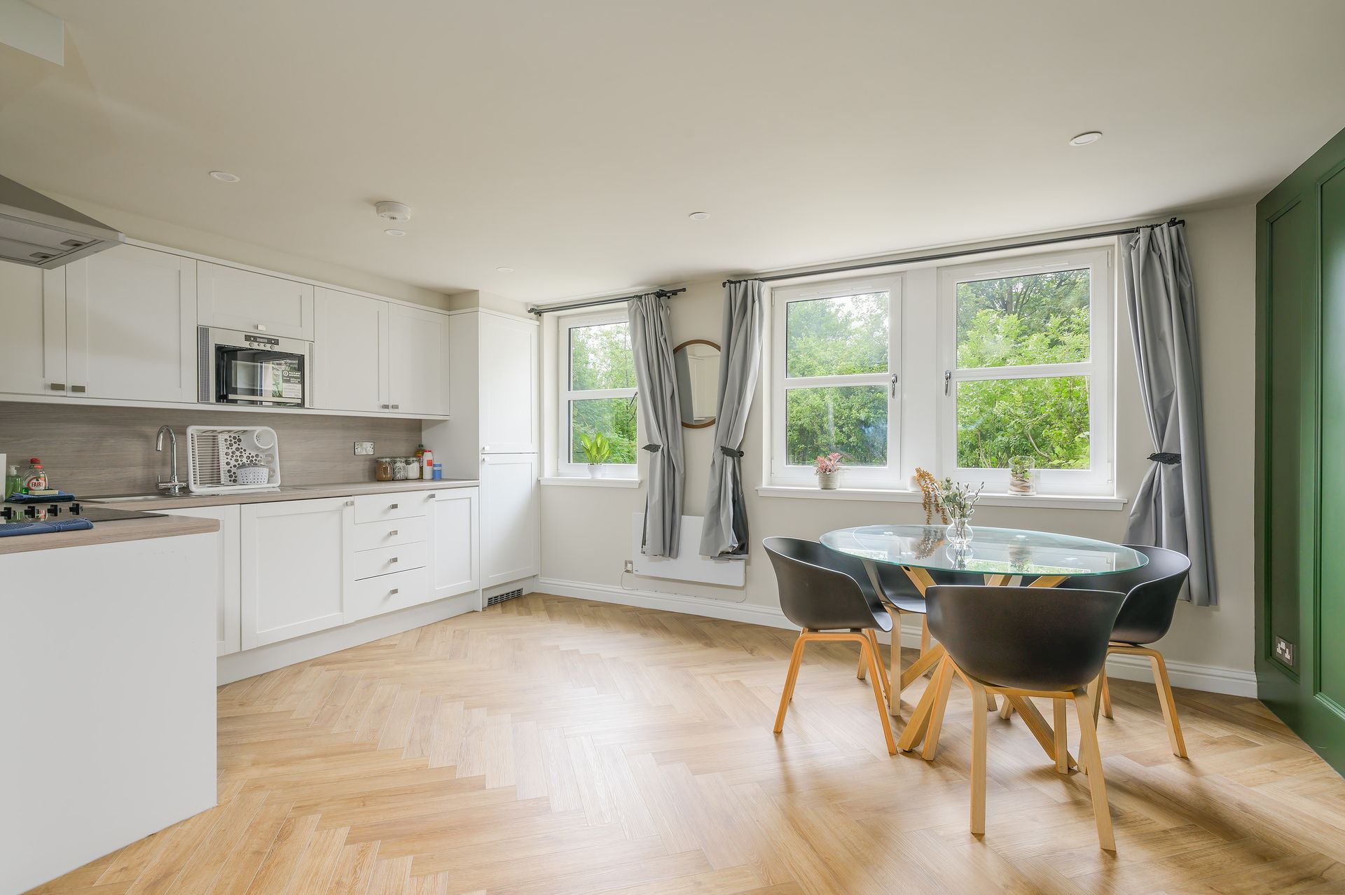 Open-plan kitchen/dining room with white cabinets, light wood flooring, a round table, and three windows with curtains.