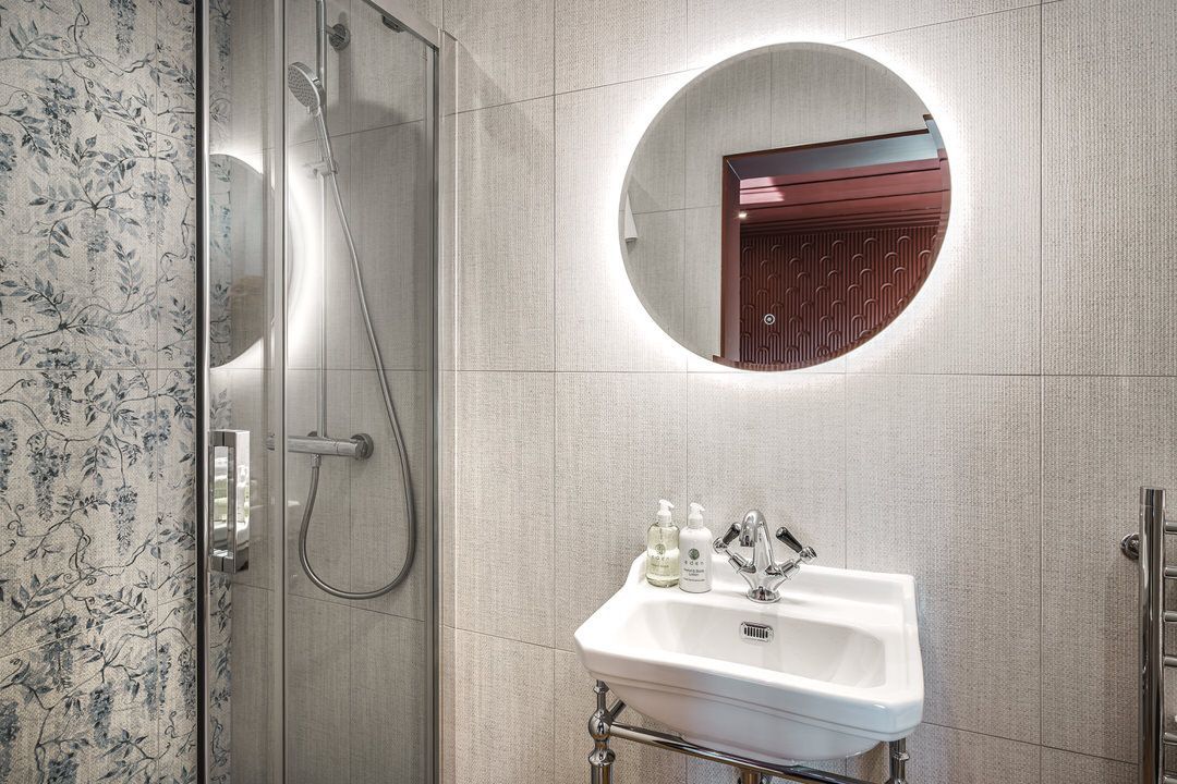 Bathroom with shower, sink, and round mirror with a lit border. Gray and white tones dominate.