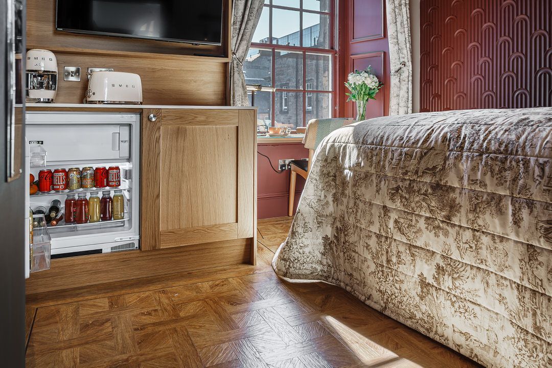 Hotel room interior with bed, built-in mini fridge stocked with drinks, and window.