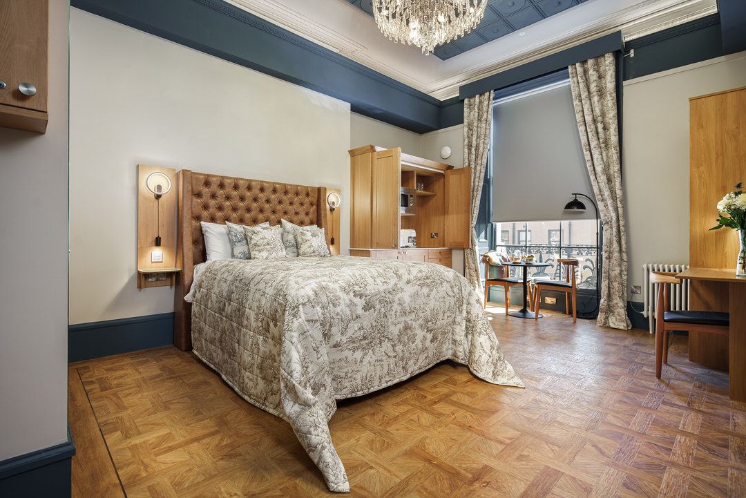 Bedroom with bed, floral bedding, chandelier, wooden floor, built-in cabinets, and large window with curtains.