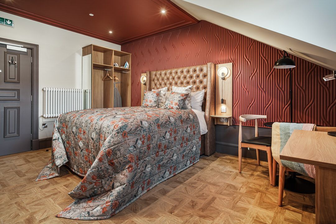 Bedroom with a bed, table, and chair. Red walls, patterned bedspread, wooden floor.