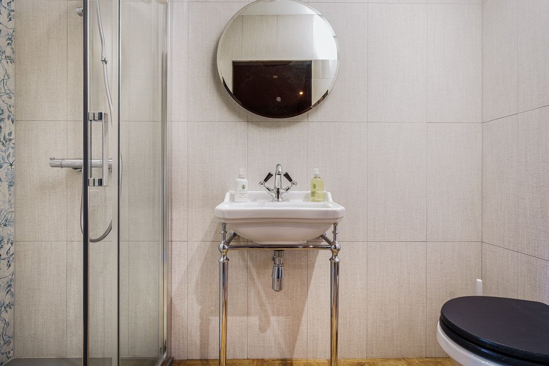 Bathroom with a white sink, round mirror, shower, and toilet. Beige tiles cover the walls.