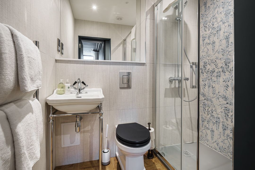 Small bathroom with a toilet, sink, and shower. White walls and wooden floor. Towels hang on the wall.