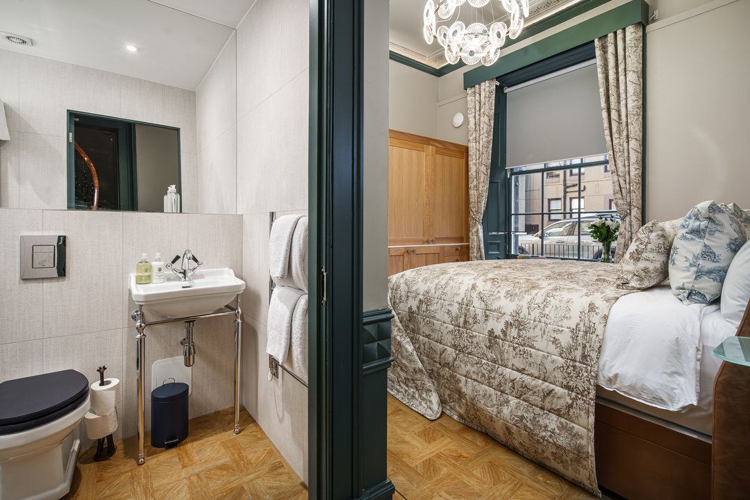 Bathroom and bedroom in a hotel suite. White tile, tan and green decor, bed with floral quilt.
