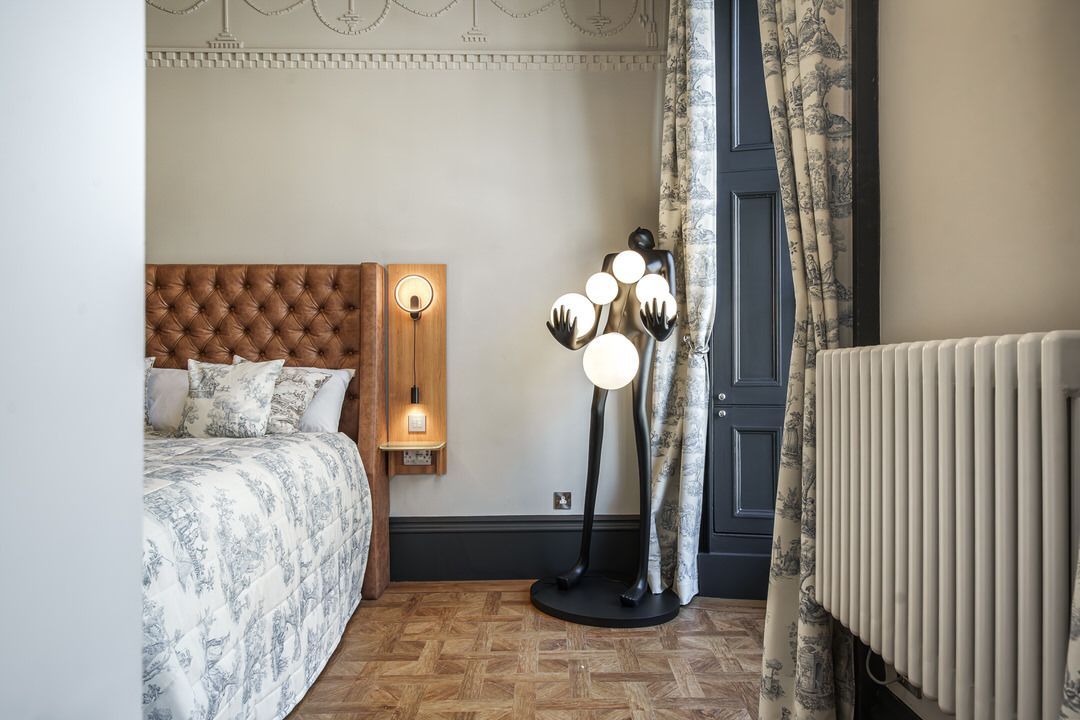 Bedroom with bed, floor lamp, patterned curtains, radiator, and wood floor.