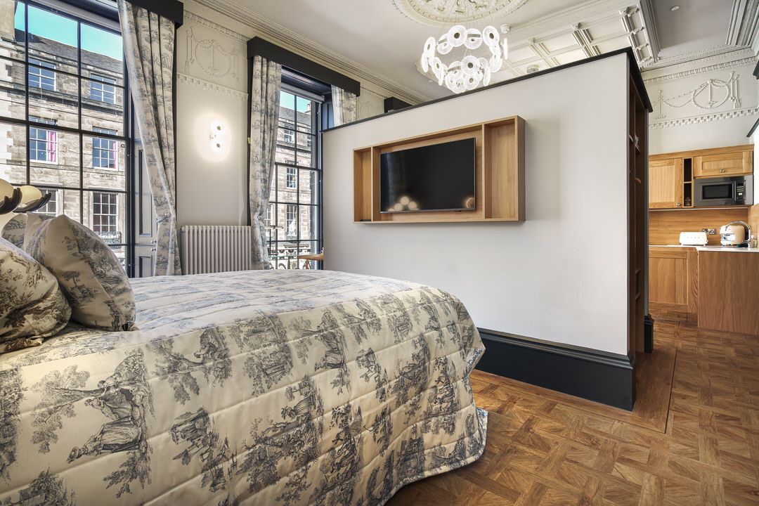 Bedroom with bed, a TV built into a wall, and a kitchenette. Sunlight streams through windows.