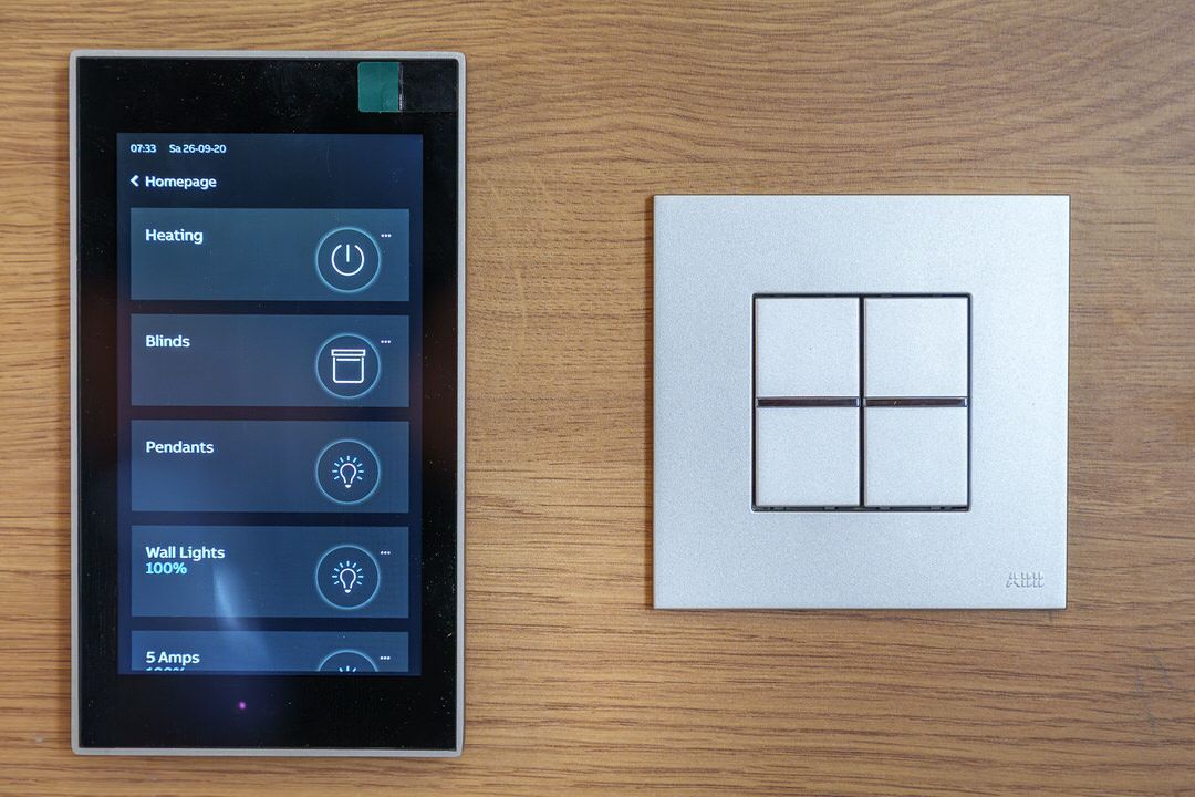 Smart home control panel and light switch on a wooden surface. The panel displays various smart home functions.