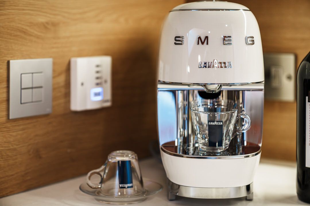 White Smeg coffee maker with glass carafe and a small cup on a counter, near wall outlets.