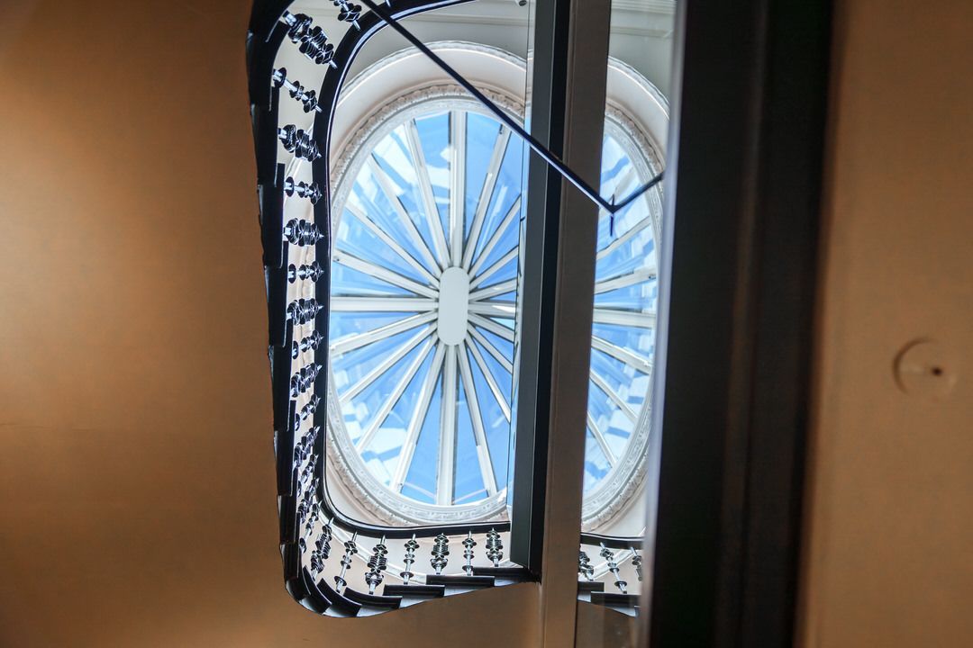 Oval-shaped window with a blue sky view, intricate white detailing, and patterned curtain.