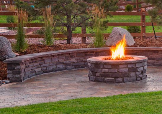 Backyard patio with stone fire pit burning beside a curved retaining wall and landscaped garden