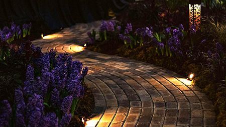 Curving brick garden path lit by warm ground lights, bordered by purple flowers at dusk