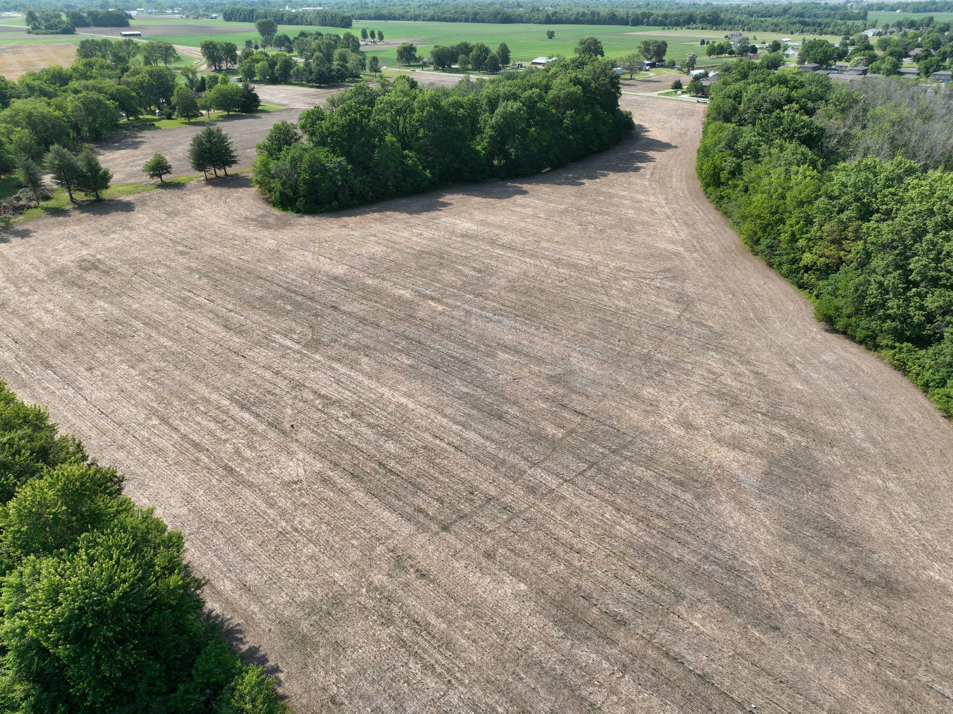 Aerial view of a plowed field bordered by green trees and farmland under clear daylight
